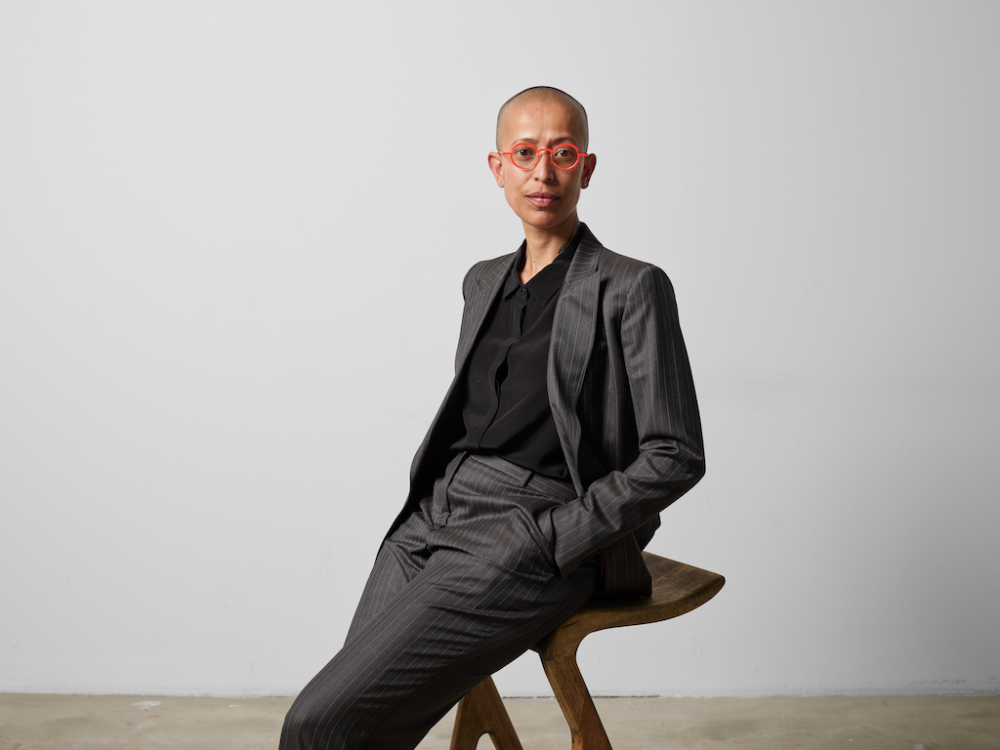 A person in a grey suit and pink spectacles sits on a backless chair.