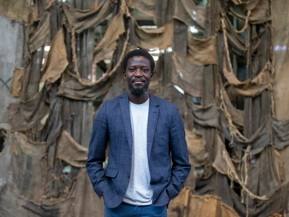 Portrait of Ibrahim Mahama standing in blazer in front of one of his jute sack sculptures.