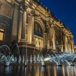 A lit-up building at twilight with a fountain in front of it.