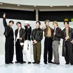 Five young Korean men in trendy clothing standing side by side on a white stage posing after a performance.