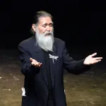 Portrait of Agosto Machado in a black blazer with his hands raised. He has a large dragonfly brooch affixed to his blazer.