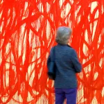 A woman seen from behind in front of a monumental painting filled with red swirls.