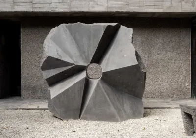 A solid looking grey sculpture that looks like a wheel with rough, slightly squared off edges and an angular surface.