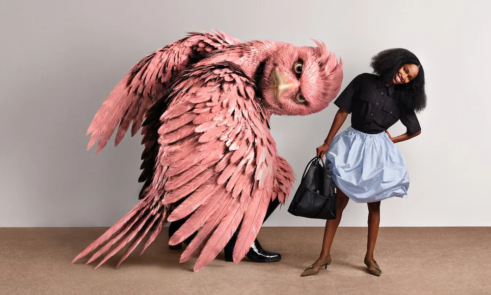 A woman in a blue skirt and black shirt standing with one arm bent and smiling. She stands next to a giant pink bird that appears to be digitally rendered.