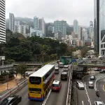 HONG KONG, CHINA - MARCH 20: Traffic moves along a busy urban road with double-decker buses and cars under overcast skies on March 20, 2026 in Hong Kong, China. Residential and commercial high-rise buildings are seen against a hillside partially covered by low clouds, reflecting the city
