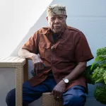 Portrait of Melvin Edwards seated at a table with one arm resting on it.
