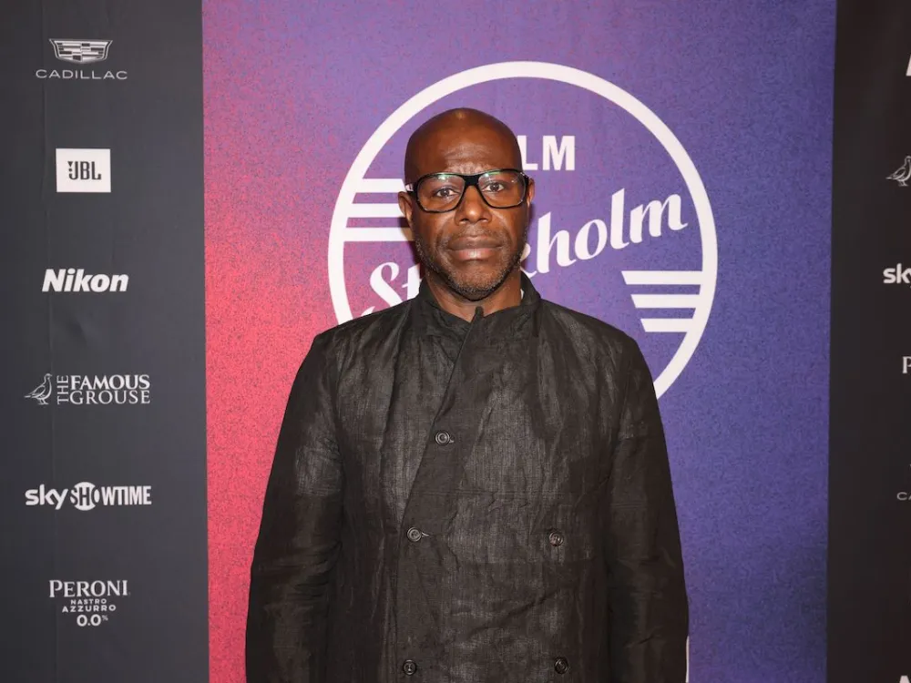 Portrait of Steve McQueen, a Black man in a black suit standing on a red carpet.