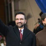 A man in a black jacket and a woman in a brown coat beaming in front of an audience.
