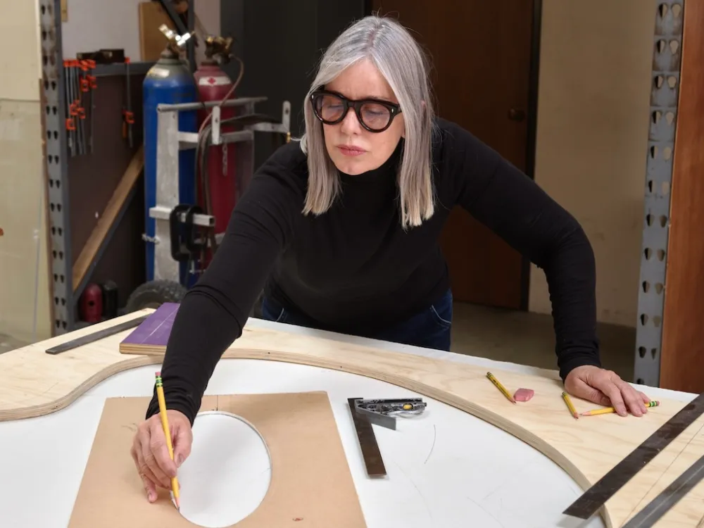 I woman with straight grey hair in black and thick black glasses draws on a large piece of paper.