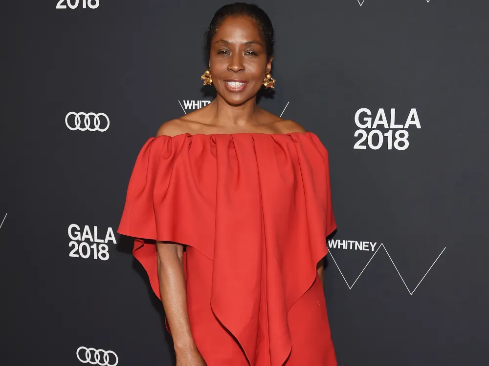 A Black woman, Lorna Simpson, attends the 2018 Whitney Gala  on May 22, 2018 at Whitney Museum of American Art in New York City. She wears a red dress and stands in front of a step-and-repeat backdrop indicating the setting is the Whitney Gala in 2018 and bears the Audi logo