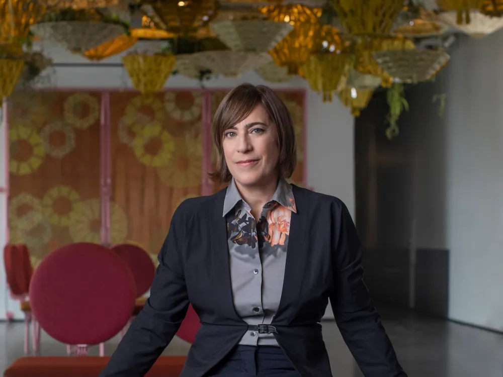A woman, Madeleine Grynsztejn, is seated on a low table with her arms spread out to her sides, looking at the camera
