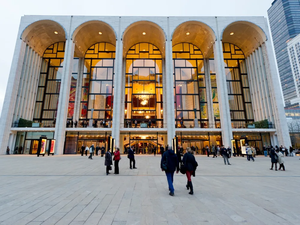 A photo shows the home of the Metropolitan Opera at Lincoln Center in New York, with a colonnade fronting several-story-high windows. Several people stand on a large plaza in front of the building.