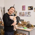 Ming Wong in his studio, holding a stuffed animal of a pig, with photos of St. Sebastian hanging on the wall.