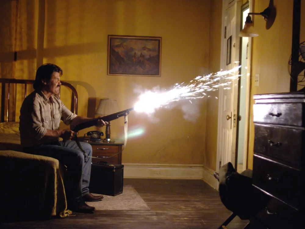 A man seated on a bed in a motel room fires a shot gun at a door that is opening.