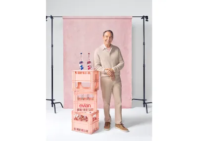 A smiling man in tan pants and a sweater standing in front of a light pink photo backdrop, with his elbow resting on a stack of light pink crates with two glass bottle resting on top