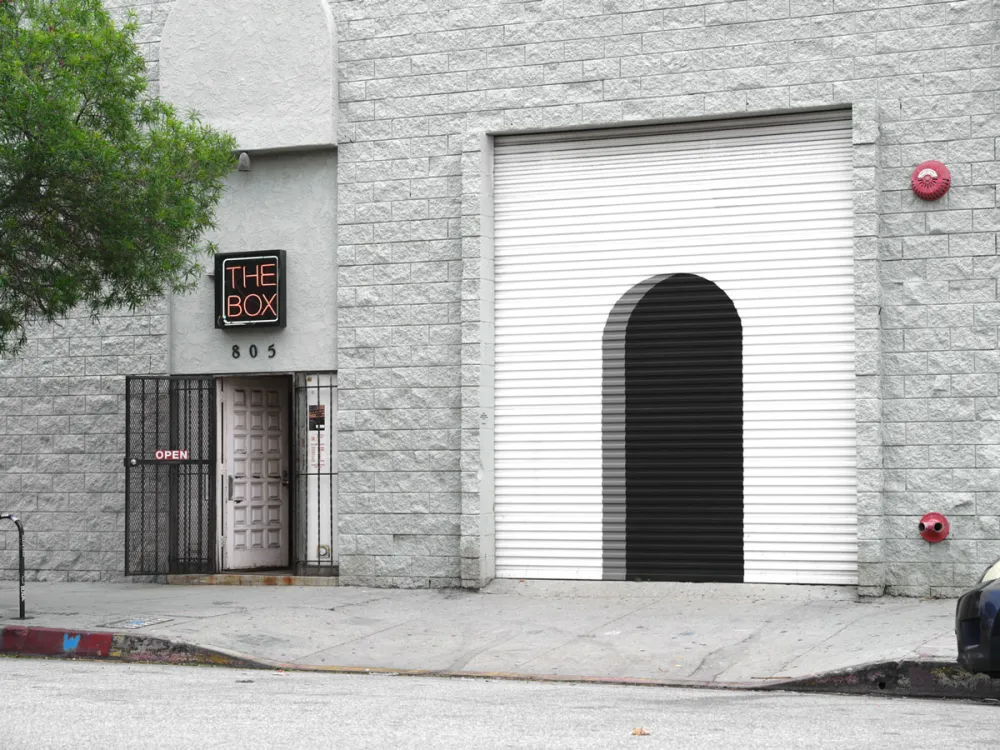 View of a gray building with a mural on the garage of an arched entryway. To the left reads a neon sign 