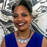 Portrait of Kymberly Pinder. She is wearing a blue top and a silver necklace and is standing in front of a black-and-white artwork.