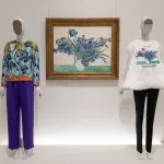 A painting of a vase with irises in it next to two mannequins, one of which wears a shirt with that same images printed on feathers.