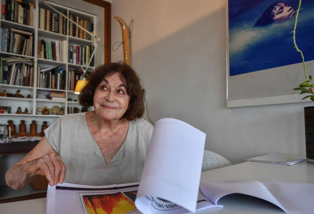 An older woman with chin-length brown hair sitting at a desk in front of tall bookcases, flipping through a large portfolio