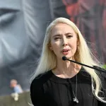 Portrait of Melissa Chiu speaking into a microphone before a large painting of a person