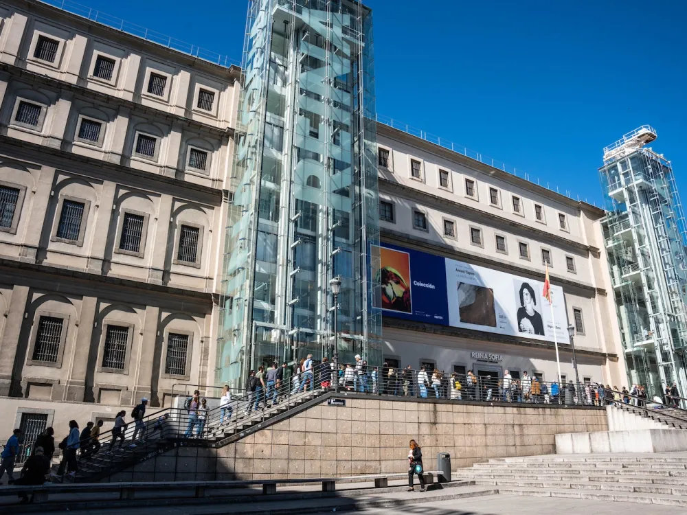 Visitors line the stairs leading to the Reina Sofía museum in Madrid, a long official looking four story building, set against a blue sky.