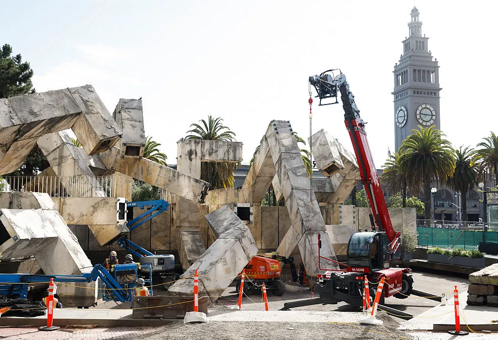 several construction cranes around a brutalist public artwork made of jumbled concrete tubes