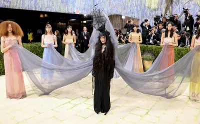 A woman in a black dress with a hat in the shape of a ship. Six other women hold grey pieces of fabric attached to it.
