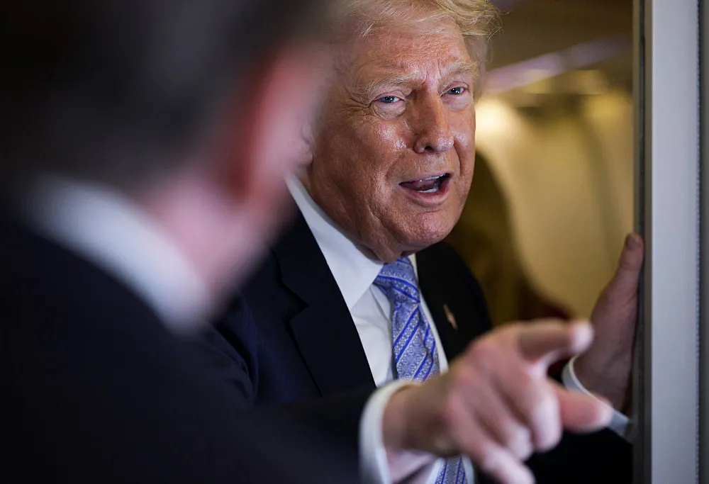 A smirking man in a dark suit and a blue tie points to something.