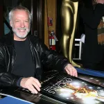A man with white hair and a close-cropped white beard smiling, sitting at a table with a poster of a gold award on the table in front of him