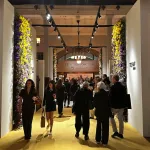 Well dressed people standing on a mustard yellow carpet in between white walls covered in flowers.