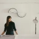 A brown haired lady in a black shirt stands behind a table. In the background on the wall is a clock that looks to have been split in half horizontally.
