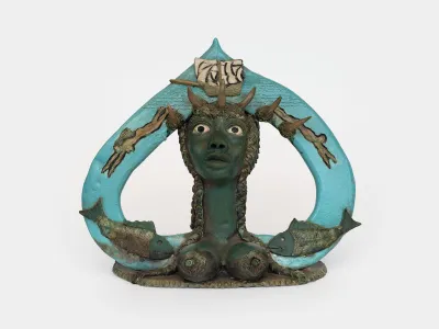 A sculpture of a female figure with her breasts bared surrounded by a blue arc. Two fish sit at either side, and a boat appears at the top.