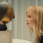 a woman with long blonde hair and bangs in profile bending down and looking at a bronze sculpture of a head on a pedestral