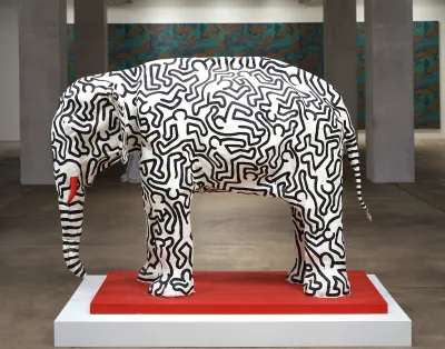 A white sculpture of an elephant covered in the outlines of leaping figures. One of its tusks is painted red, and the base is also red.