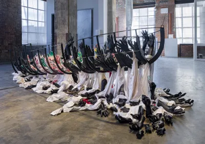 A sculpture resembling the armature of a ship held up by many black hands, some of which are sprawled out on the floor beneath amid white fabric.