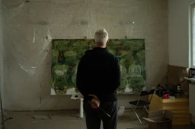 A man, with his back to the camera, looks at a painting in-progress in his studio. He holds a paintbrush. 