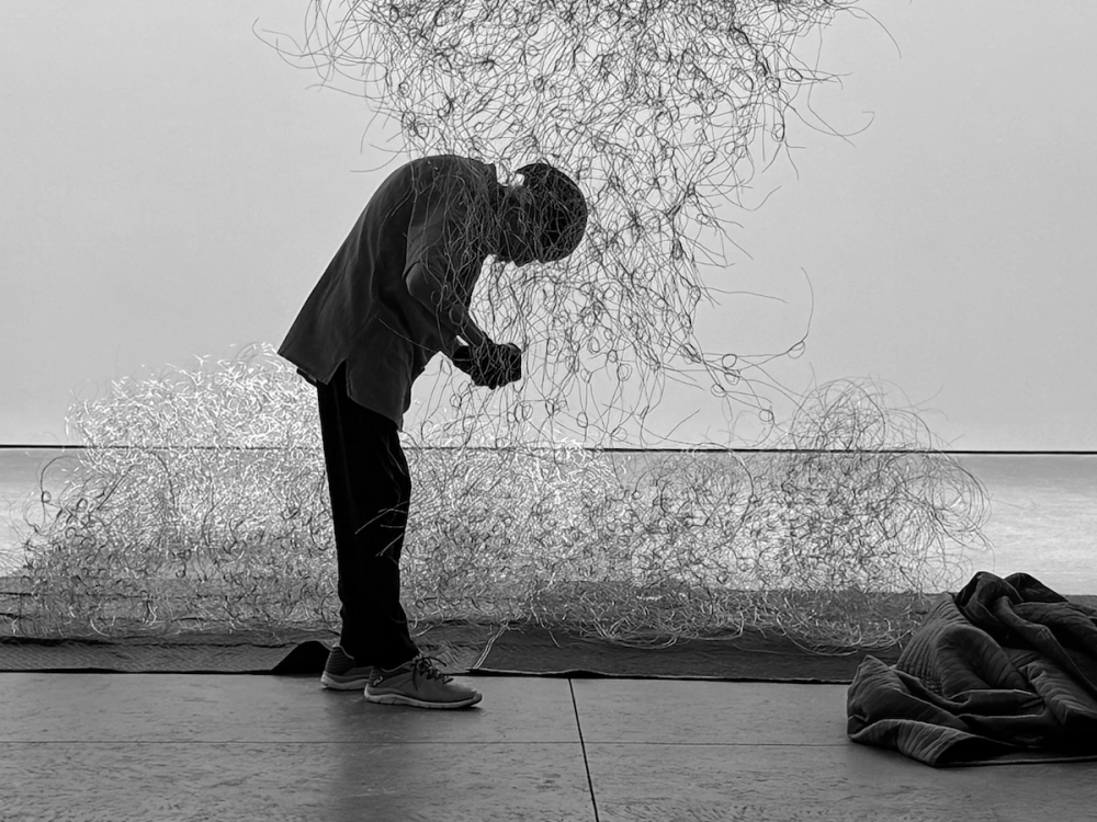 A man crouched beneath a suspended wire sculpture.