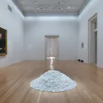 A gallery with a pile of white candies on its floor. A painted portrait hangs on one wall, and a hanging string of lights can be seen in an adjacent space.