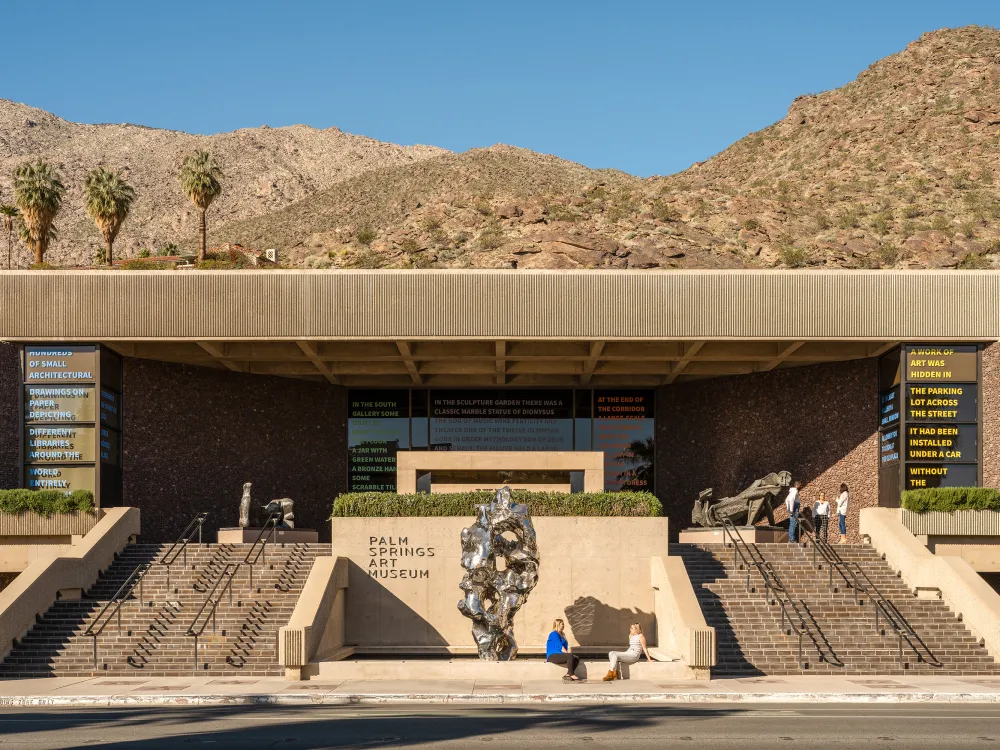 A photo shows the facade of the Palm Springs Art Museum, a low-slung modernist structure, with a silver sculpture and several visitors