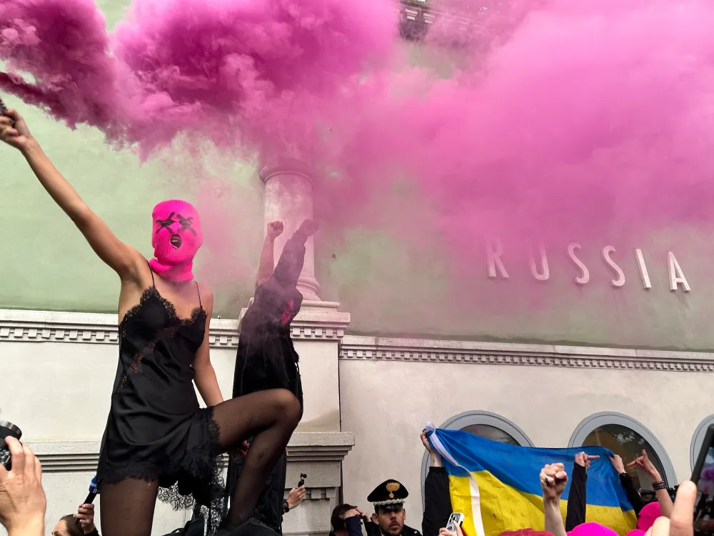 A woman in a black slip and a pink balaclava holds up a flare making pink smoke in front of a wall that reads 