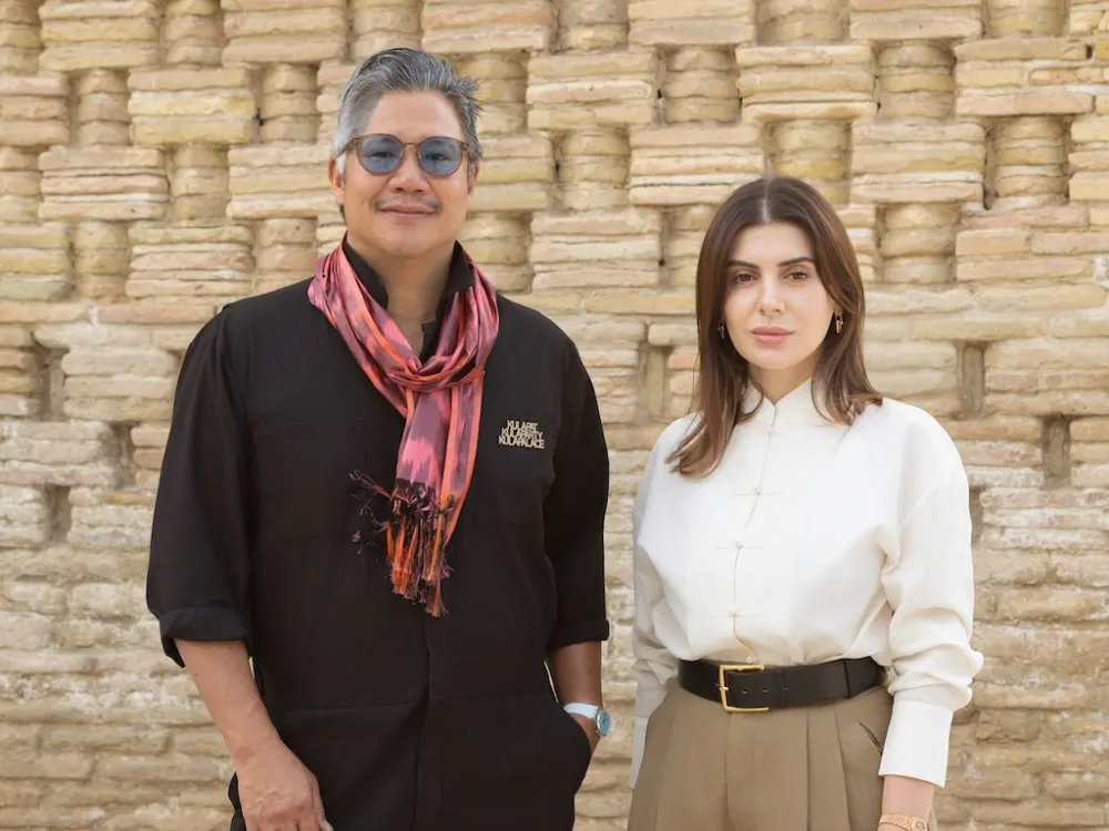 A man dressed in black with a reddish scarf around his neck stands next to a woman with a white shirt, kahki pleated pants, and medium length brown hair.