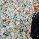 A man in a dark suit stands next to an abstract painting.
