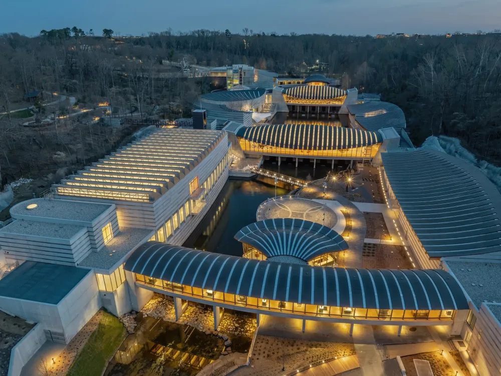 An aerial photo shows the Crystal Bridges Museum of American art at dusk, its galleries lit up from the interior, in a wooded landscape