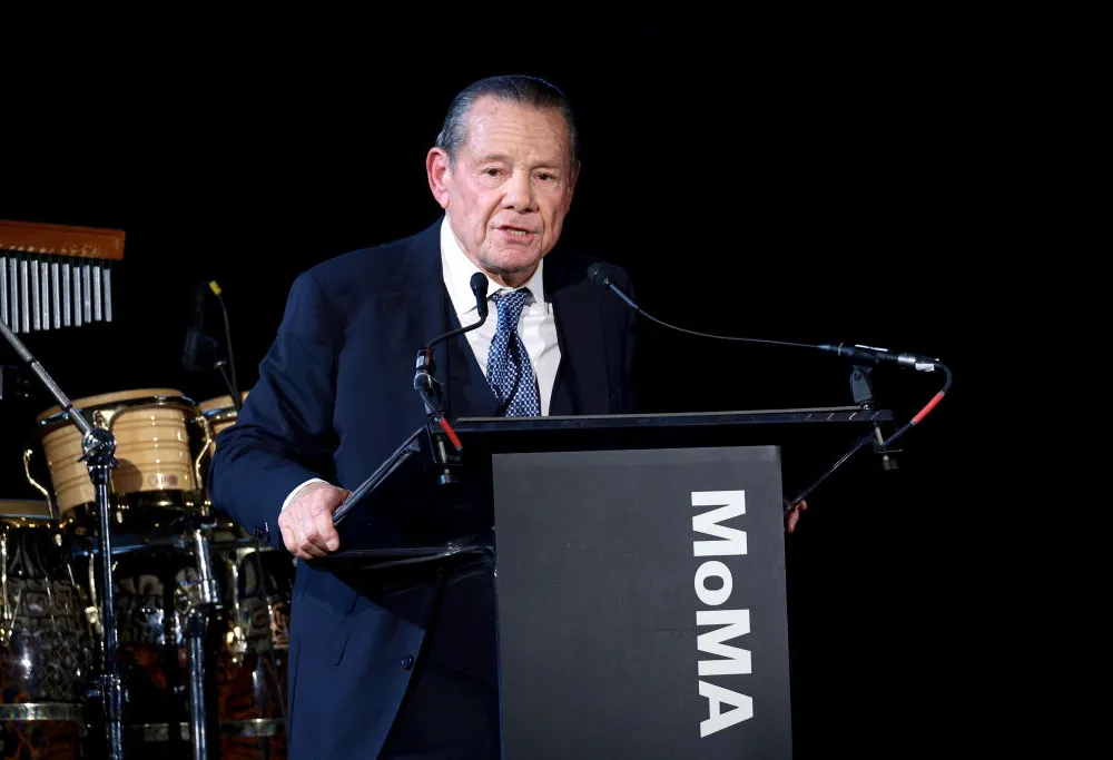 A man in a dark blue suit, white shirt, and tie standing on a stage behind a lectern with 