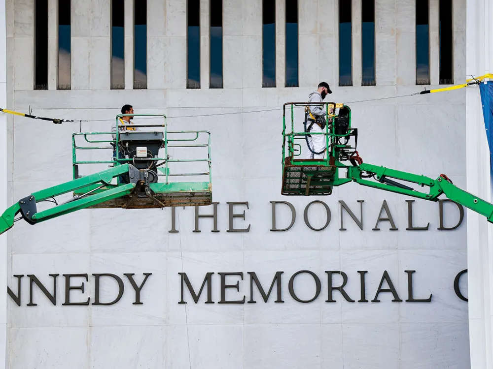 A stone facade bearing black letters and flanked by green cranes carrying men.