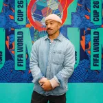 A man in a denim shirt and pink beanie poses in front of a blue gradiented large graphic design poster.