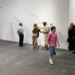 several people, including a man in a khaki jumpsuit standing near a while wall, in a gallery booth space at an art fair