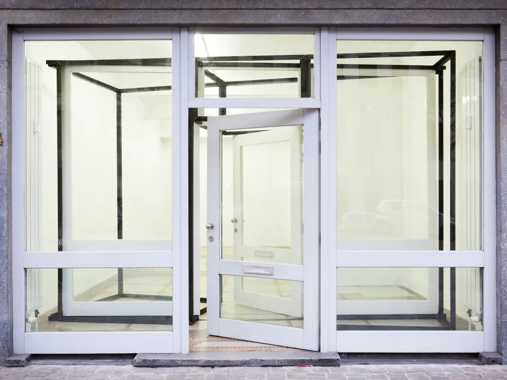 View of a gallery with two large glass windows flanking a glass door. Inside is a clear, metal structure.