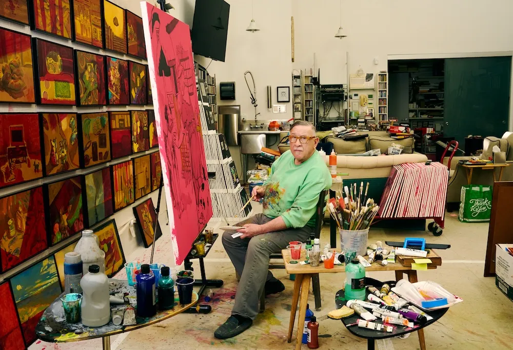 A gentleman in a paint smeared green sweatshirt sits in the middle of a painting studio while working on a canvas.