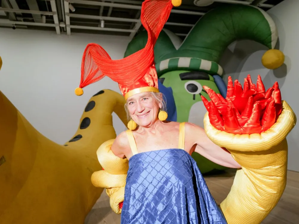 A woman in a blue dress with a big red hat and big puffy yellow earrings. She stands next to a large inflatable showing a green face and a long yellow trumpet.